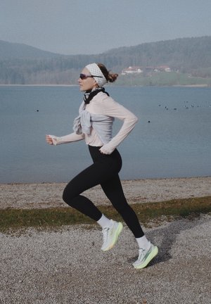 Mujer con mallas negras y zapatillas blancas corriendo por un camino de grava junto a un lago, con colinas y casas al fondo.