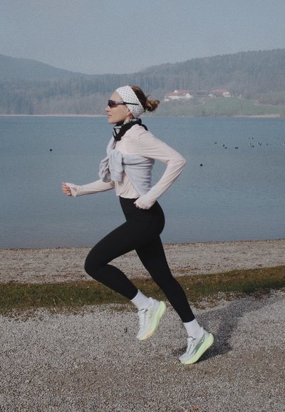 Mujer con mallas negras y zapatillas blancas corriendo por un camino de grava junto a un lago, con colinas y casas al fondo.