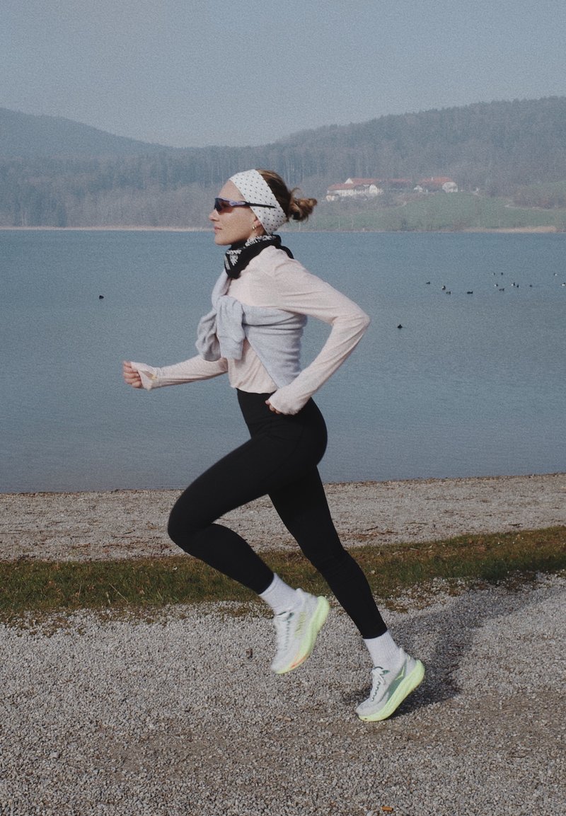Nainen mustissa legginsseissä ja valkoisissa lenkkareissa juoksee soratiellä järven rannalla, taustalla mäkiä ja taloja.