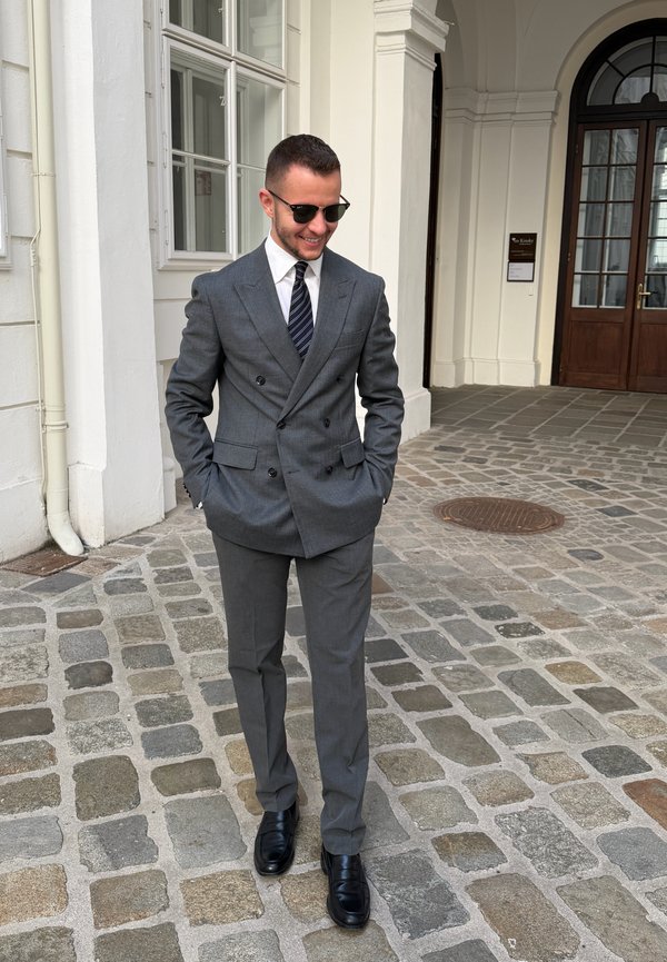 Un hombre sonriente con traje gris, corbata a rayas y gafas de sol está de pie en un camino empedrado, frente a un edificio blanco.
