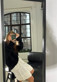 Giovane donna in maglione nero e gonna bianca seduta su uno sgabello, mentre fa un selfie allo specchio con il telefono in una stanza moderna con una grande finestra.