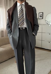 Costume gris avec une finition légèrement texturée, pull marron drapé sur les épaules, cravate rayée dans des tons beige et marron, et ceinture avec boucle argentée.