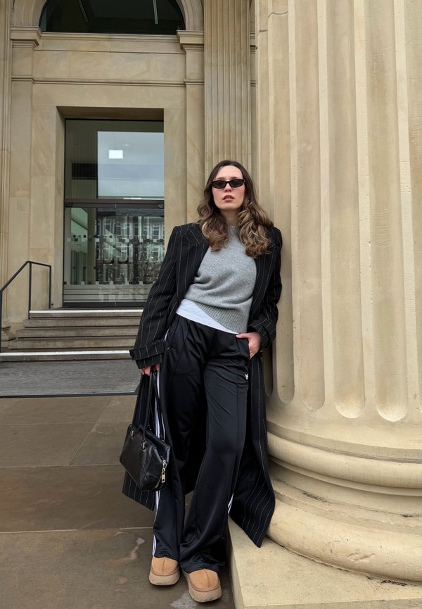 Woman wearing a black pinstripe coat, grey sweater, black wide trousers, sunglasses, and tan shoes, leaning against a large stone column outside a building.