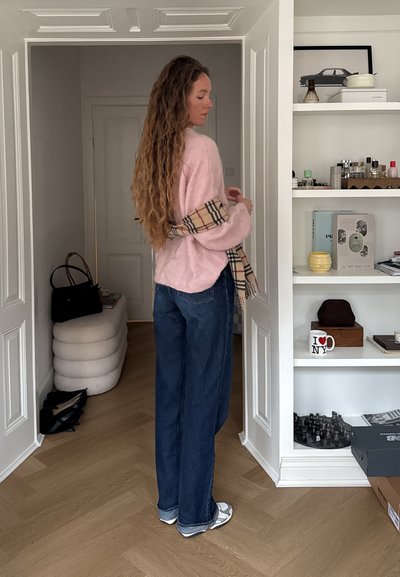 Mujer con cabello largo y rizado, vestida con un suéter rosa y jeans azules, de pie junto a una estantería blanca con decoración y botellas de perfume.
