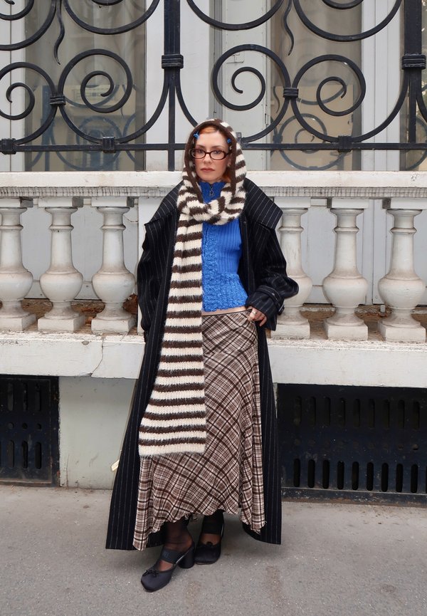 Une femme pose devant une élégante rambarde en fer forgé. Elle porte un long manteau à fines rayures, un pull bleu, une écharpe rayée, une jupe à carreaux et des lunettes.