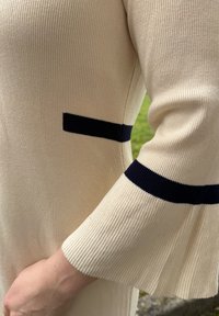 Sweter dziany w kolorze kremowym z ciemnoniebieskimi paskami na mankietach i kieszeni, charakteryzujący się prążkowaną fakturą i rozszerzanym fasonem rękawów.