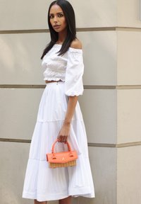 Frau mit langen, dunklen Haaren, die ein weißes, schulterfreies Oberteil und einen plissierten Rock trägt und eine kleine orangefarbene Handtasche aus Rattan hält.