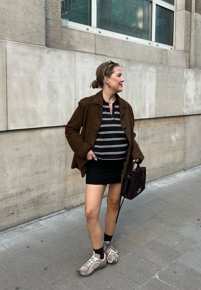Mujer vestida con una chaqueta marrón, una camiseta de rayas, una falda negra y zapatillas, de pie en una acera urbana sosteniendo un bolso de mano tejido oscuro a su lado.