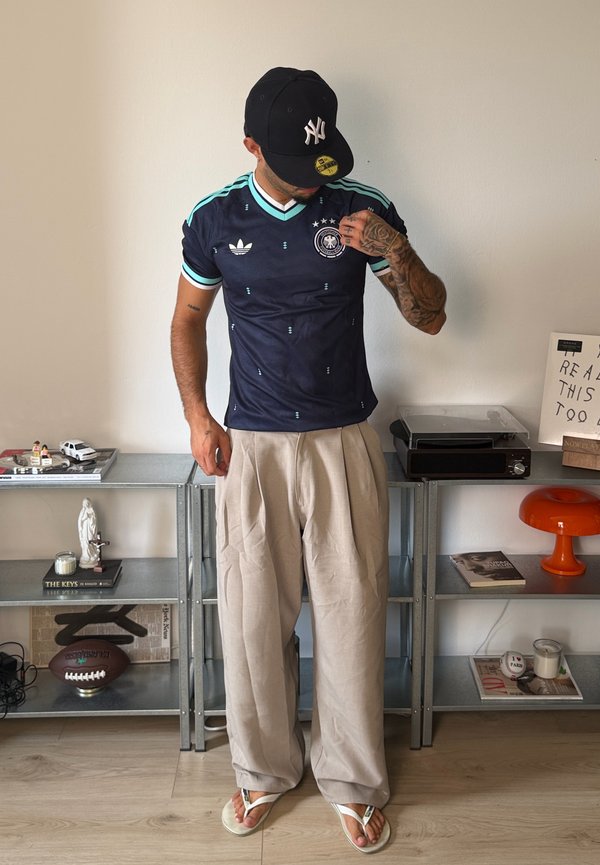 Hombre con una camiseta de fútbol de Alemania azul oscuro, pantalones anchos beige, chanclas blancas y una gorra de béisbol negra mirando hacia abajo, de pie en una habitación.