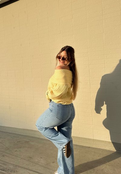 Mujer con suéter amarillo de hombros descubiertos y jeans azules posa con una pierna levantada, sonriendo y usando gafas de sol frente a una pared beige bajo la luz del sol.