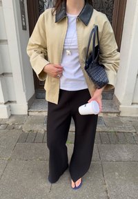 Jachetă oversized bej cu guler din piele neagră, tricou alb, pantaloni largi negri și sandale albastre tip flip-flop. Ținând o cană albă de cafea.