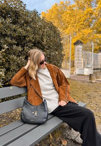 Casaco oversized de camurça castanha com piping preto, suéter cinza claro, calças de perna larga pretas e uma bolsa de couro preta num banco de parque.