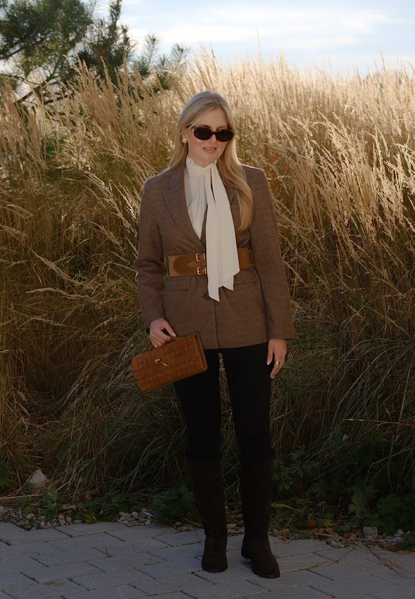 Une femme portant des lunettes de soleil, un blazer brun, une écharpe blanche et des bottes tient un sac à main brun près de hautes herbes.