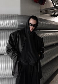 Svart läderjacka över en svart luvtröja, kombinerad med mörka solglasögon. Bakgrunden har silverfärgade metallkanaler.