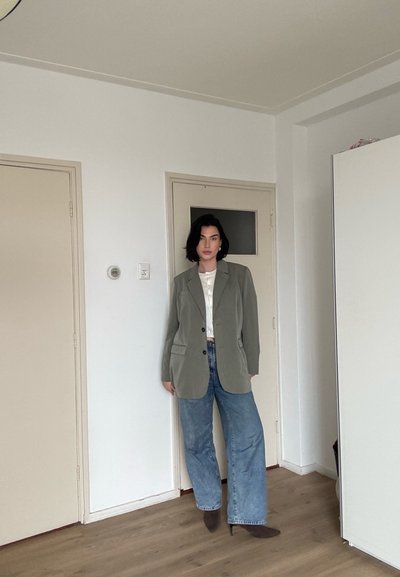 Mujer con cabello corto y oscuro que lleva una chaqueta oliva, blusa blanca, jeans de pierna ancha y botas marrones con punta, de pie en el interior junto a puertas cerradas.