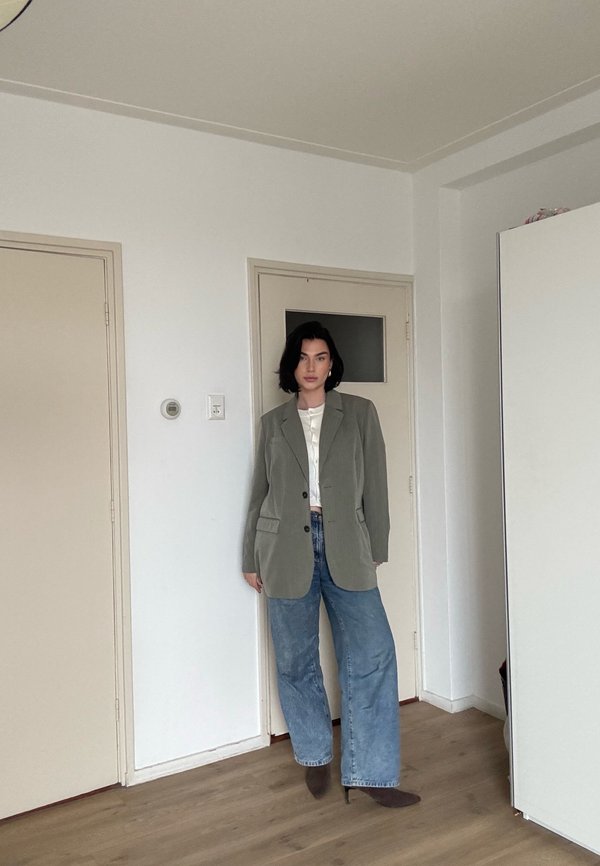 Žena s krátkými tmavými vlasy má na sobě olivově zelené sako, bílý top, široké džíny a hnědé špičaté boty, stojí uvnitř u zavřených dveří.