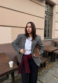 Grey tweed blazer over a white t-shirt, black trousers, and a maroon scarf tied at the waist; holding a brown bag; paper cup nearby.