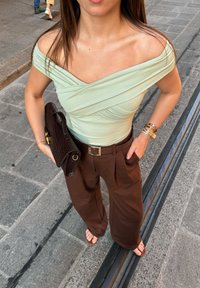 Ženska v svetlo zeleni bluzi brez naramnic, rjavih širokih hlačah, drži temno rjav teksturiran clutch, stoji na tlakovani ulici.
