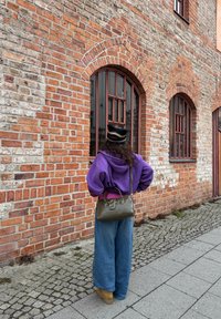 Personne portant un sweat à capuche violet, un jean large et un chapeau noir, debout sur un trottoir pavé, faisant face à un bâtiment en briques avec des fenêtres cintrées.