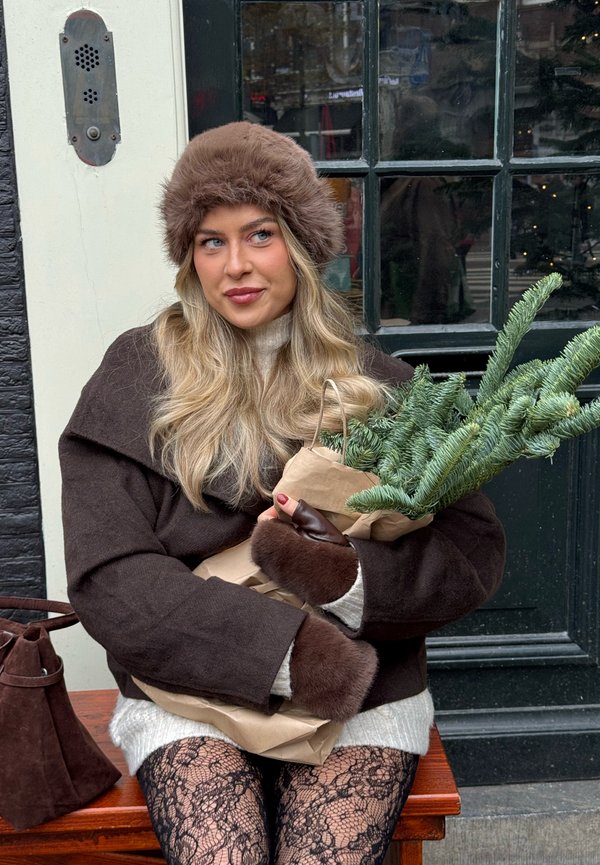 Abrigo de lana marrón con ribete de piel, suéter de punto crema, medias negras de encaje, sosteniendo un ramo de pino envuelto en papel kraft, sentado junto a una puerta.