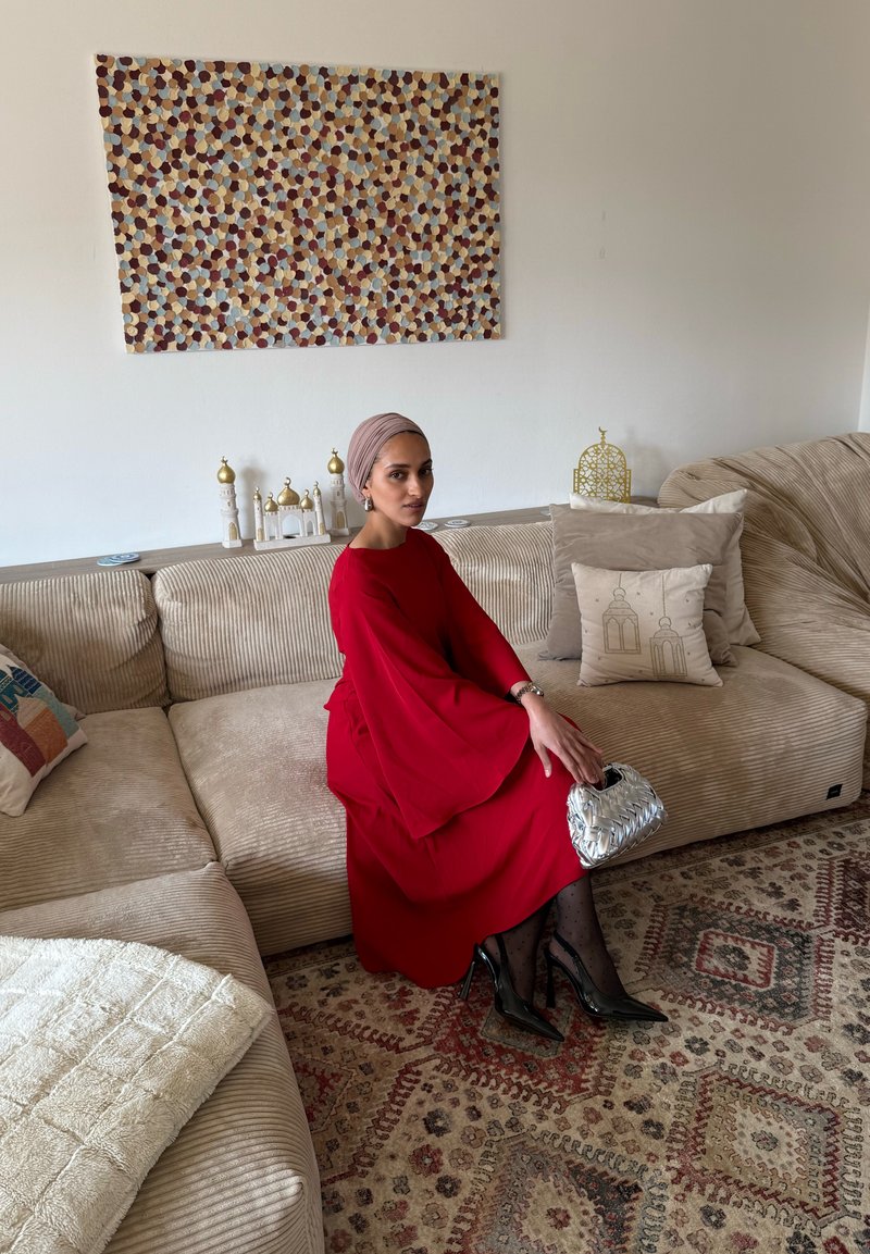 Frau in rotem Kleid und Kopftuch sitzt auf einer beigen Eckcouch, hält eine silberne Handtasche, dekorative Kissen und gemusterter Teppich sind sichtbar.