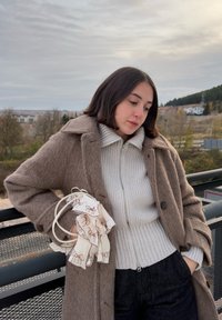 Hnedý vlnený kabát s veľkým golierom cez svetlosivý rebrovaný sveter so zipsom. Drží vzorovanú látkovú taštičku. Tmavé nohavice. Prírodné pozadie.