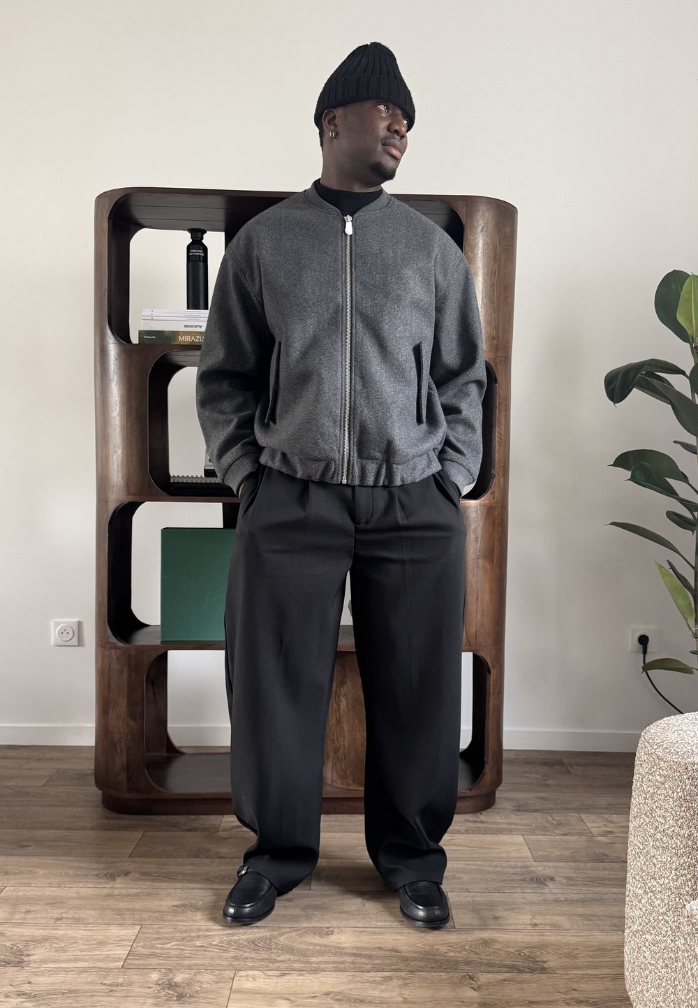 Homme portant un bonnet noir, une veste grise zippée, un pantalon noir et des chaussures noires, debout devant une étagère en bois et une plante dans une pièce.