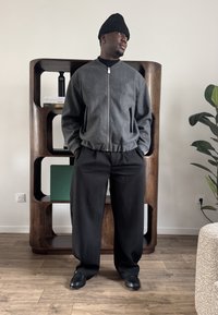 Homme portant un bonnet noir, une veste grise zippée, un pantalon noir et des chaussures noires, debout devant une étagère en bois et une plante dans une pièce.