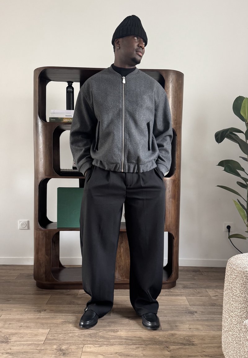 Homme portant un bonnet noir, une veste grise zippée, un pantalon noir et des chaussures noires, debout devant une étagère en bois et une plante dans une pièce.