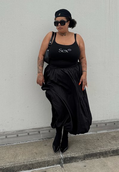 Top negro sin mangas con gráfico de "Sex", falda fluida negra, medias negras y tacones de punta. Los accesorios incluyen gafas de sol grandes y una gorra.