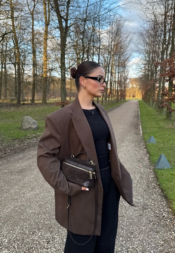 Brun överdimensionerad blazer i strukturerad tyg, i kombination med en svart topp och högmidjade byxor. Håller en liten mörk handväska med guld detaljer.