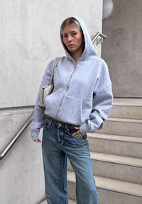 Harmaa vetoketjullinen huppari, jossa on suuri huppu ja sivutaskut, yhdistettynä löysiin sinisiin farkkuihin. Tukevat valkoiset olkalaukku, jossa on minimaalista varaosaa.