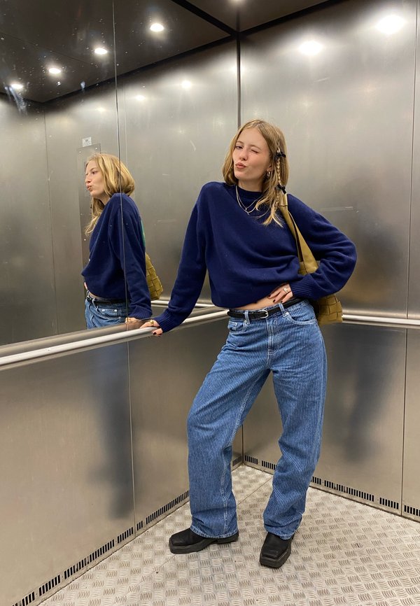 Mujer joven con suéter azul y jeans guiña un ojo y posa con una mano en la cadera dentro de un ascensor de metal con paredes espejadas.