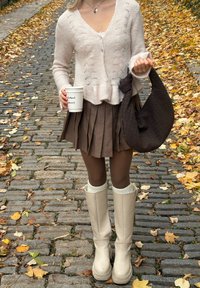 Lys beige, strikket cardigan, brun plisseret nederdel, cremede knæhøje støvler, tekstureret brun taske, holder en hvid kop, efterårsblade på brosten.