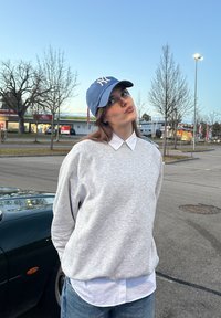 Sweat-shirt gris à encolure arrondie porté sur une chemise blanche à col, associé à une casquette de baseball bleu clair avec un logo.