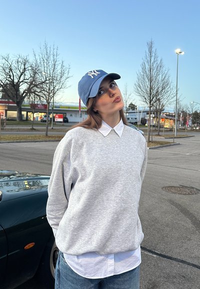 Sudadera gris con cuello redondeado usada sobre una camisa blanca con cuello, combinada con una gorra de béisbol azul claro con un logotipo.
