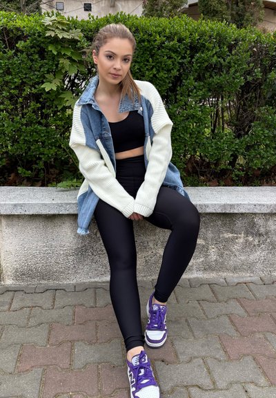 Chaqueta oversize de mezclilla y crema sobre un top corto negro, combinada con leggins negros y zapatillas moradas de New Balance, sentada en un banco de piedra.