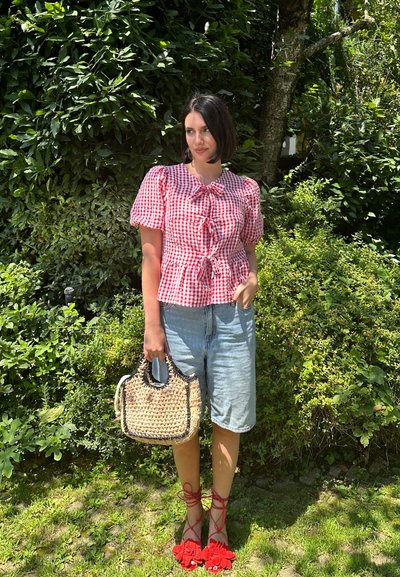 Mujer con blusa de cuadros rojos, shorts de mezclilla, sandalias rojas de encaje, sosteniendo una bolsa de paja tejida, de pie frente a un denso follaje de jardín verde.