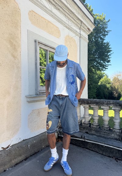 Hombre con una camisa de encaje azul, una camiseta blanca, pantalones cortos de mezclilla con parches, zapatillas azules y una gorra azul claro de pie en una terraza cerca de un edificio beige.