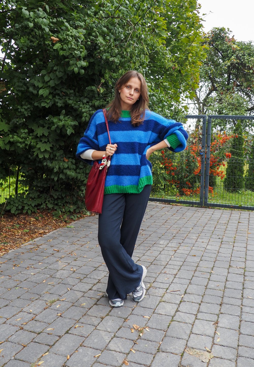 Kobieta w niebiesko-zieloną w paski swetrze stoi na wybrukowanej ścieżce obok zieleni, trzymając czerwoną torbę na ramię i nosząc sneakersy.