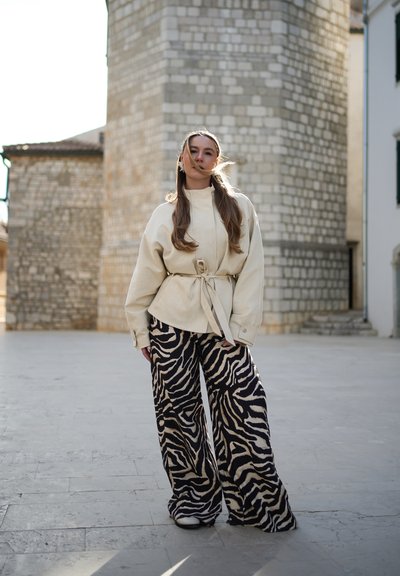 Joven mujer con cabello largo está de pie al aire libre junto a un edificio de piedra, vistiendo una chaqueta beige con cinturón y pantalones anchos con estampado de cebra en blanco y negro.