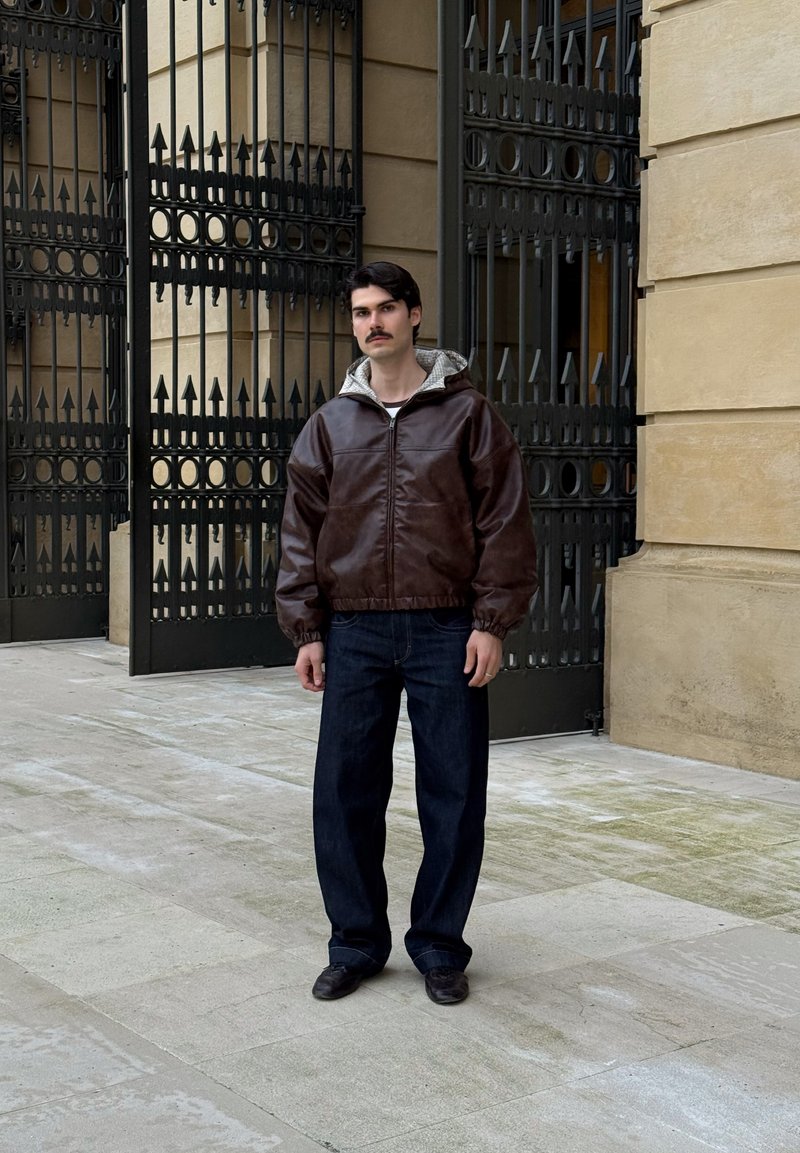 Homme aux cheveux foncés et moustache, portant une veste en cuir marron, un jean foncé et des chaussures noires, se tient sur un pavé en pierre près de grandes grilles en fer noir.