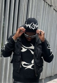 Fekete pulóver fehér absztrakt mintával, egy fekete ujjatlan dzseki alatt. Fekete sapka fehér mintával, napszemüveg, kezek a kalap igazításán.