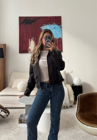 Žena s dugom kosom snima selfie u ogledalu, noseći crnu jaknu, bijelu majicu i plave traperice u modernom dnevnom boravku s apstraktnom slikom.