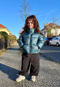 Jeune personne aux cheveux longs portant une doudoune turquoise et un pantalon noir, debout sur un trottoir dans un quartier résidentiel.