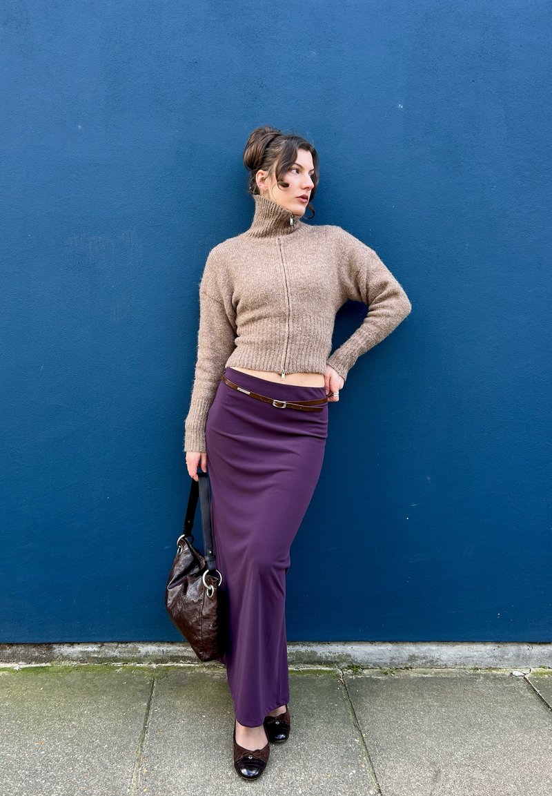 Femme debout contre un mur bleu portant un pull à col roulé beige, une longue jupe violette avec ceinture, tenant un sac à main marron, et des chaussures noires.