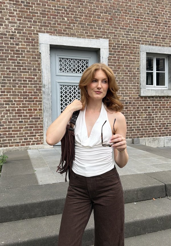 Jeune femme aux cheveux roux portant un haut blanc sans manches et un pantalon foncé, tenant des lunettes de soleil et un sac marron à franges près de l'entrée d'un bâtiment en briques.