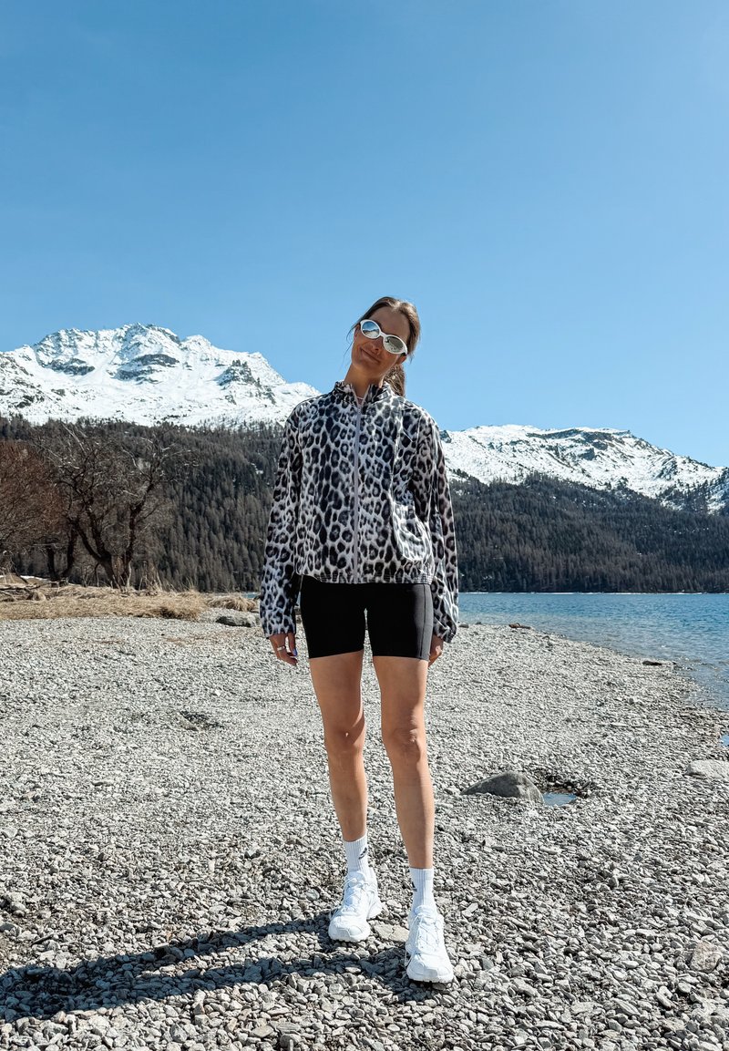 Femme portant des lunettes de soleil, une veste à motif léopard, un short noir et des baskets blanches, debout sur un rivage rocailleux avec des montagnes enneigées et un lac en arrière-plan.