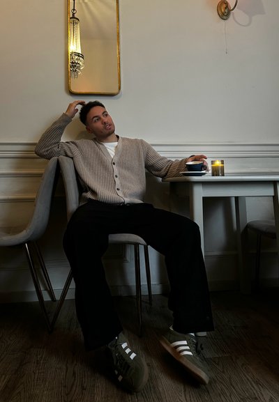 Cárdigan beige de canalé, camisa blanca y pantalones anchos negros. Zapatillas verdes con rayas blancas. Mesa de madera, sillas suaves grises y una vela.