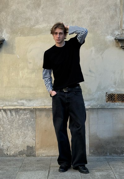 Joven con camisa de rayas de manga larga debajo de una camiseta negra, pantalones anchos negros y zapatos de cuero, de pie contra una pared texturizada beige.